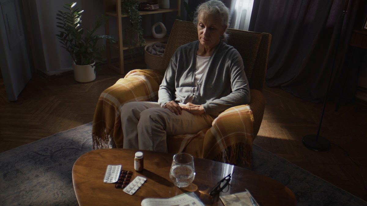 Senior woman sitting in an armchair at home, looking down at her medication on the coffee table, a pill bottle and blister packs in front of her